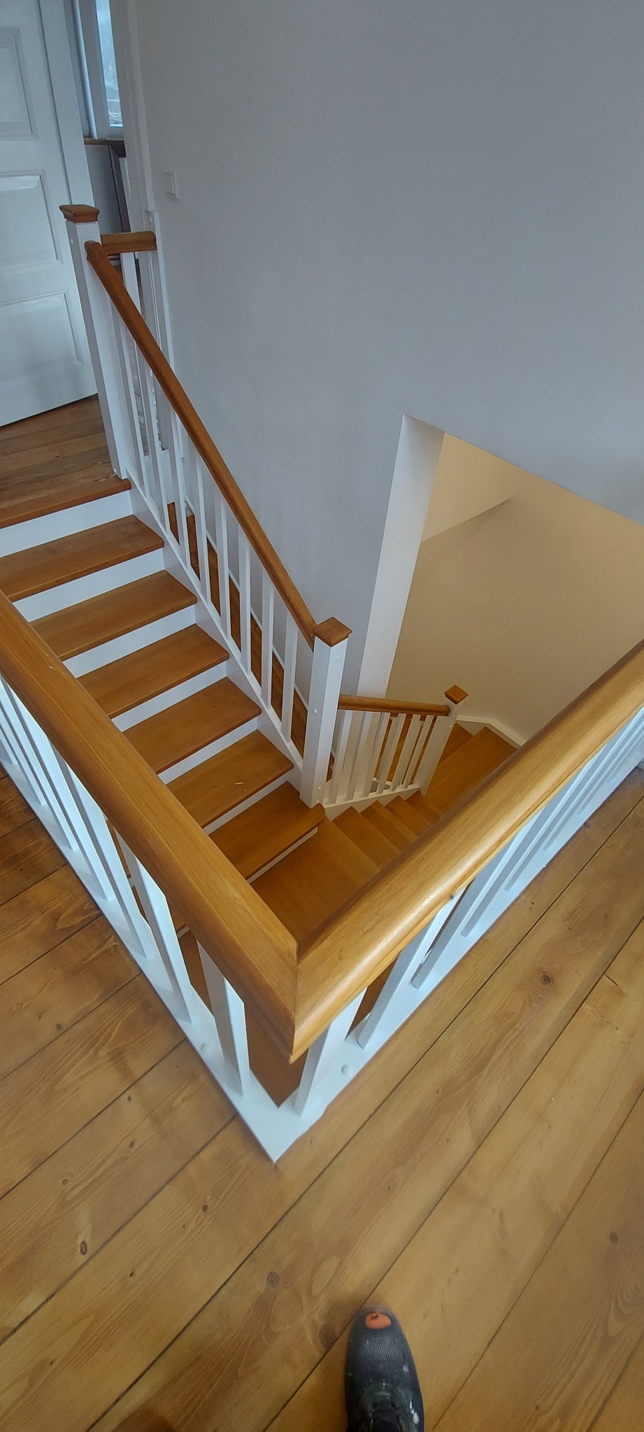 Widok z góry na odnowione schody z drewnianymi stopniami i białymi balustradami, z fragmentem drewnianej podłogi w dolnej części kadru i butem w lewym dolnym rogu.