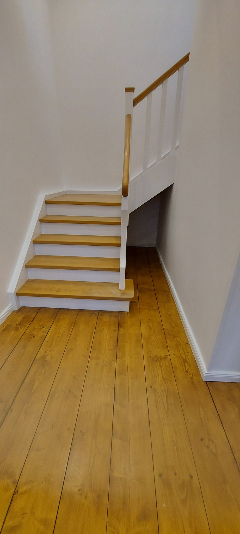 Jasne drewniane schody z białymi podstopnicami i balustradą, prowadzące na podłogę z desek w ciepłym odcieniu brązu, widok z dołu na fragment klatki schodowej.