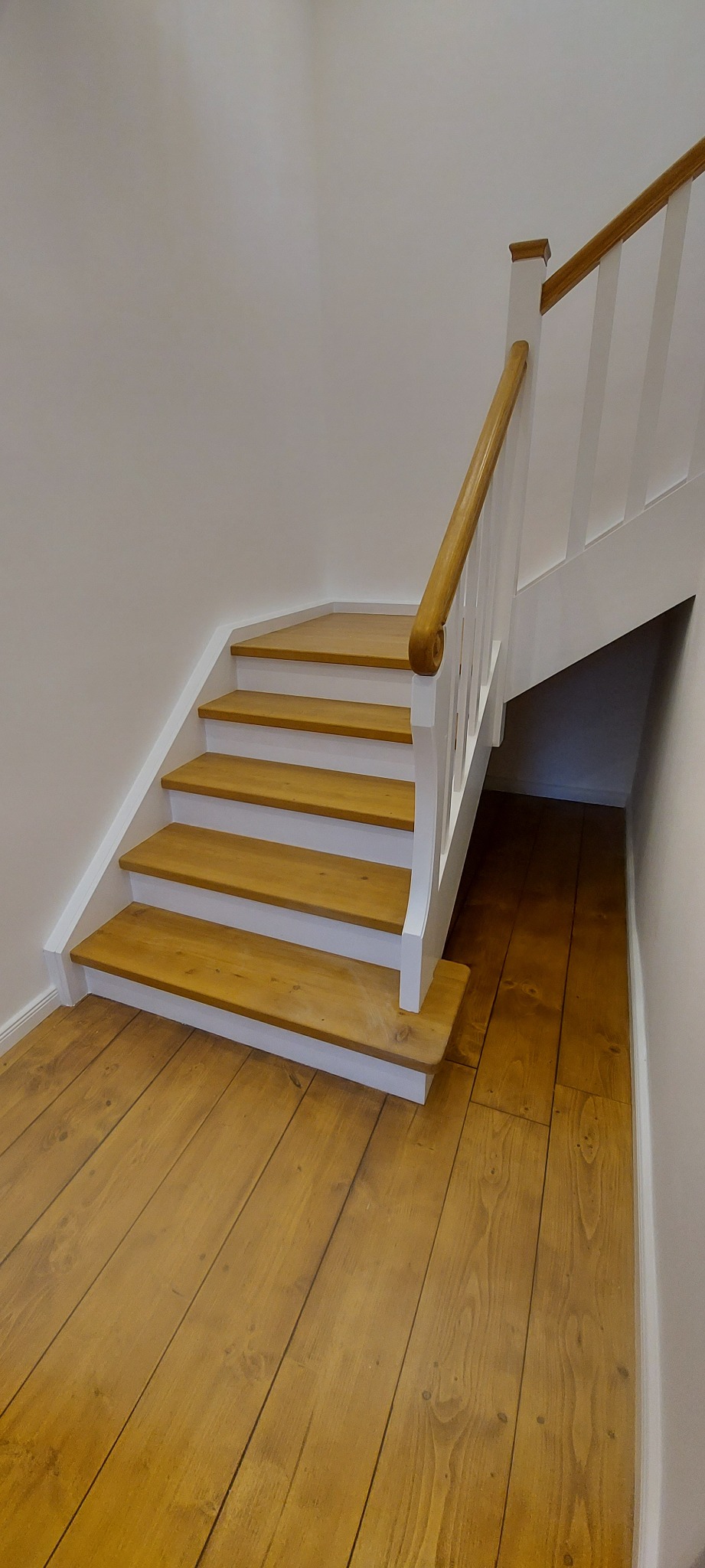 Odnowione drewniane schody z białymi podstopnicami i balustradą w Lubinie, prowadzące do ciemniejszej przestrzeni pod schodami, z widoczną drewnianą podłogą z wyraźnym usłojeniem.