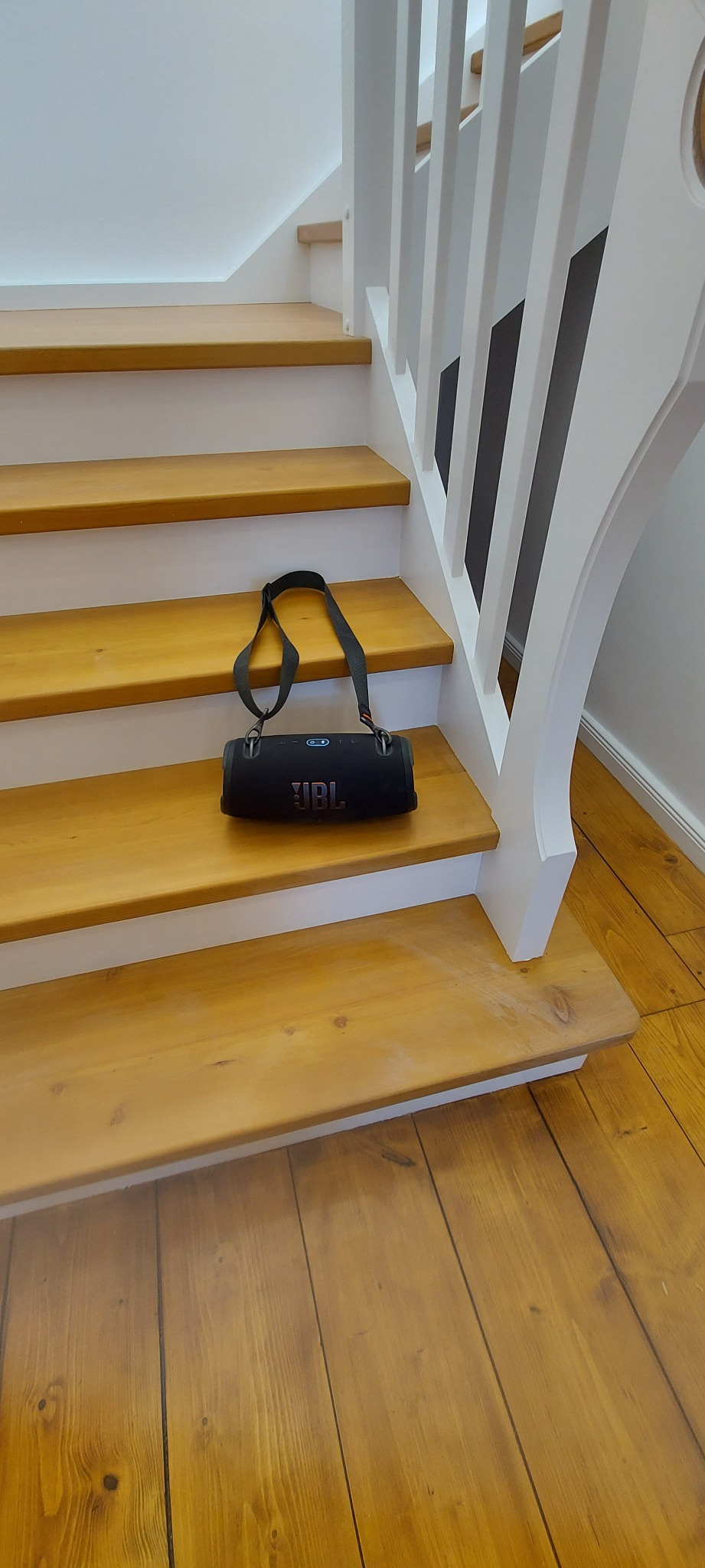 Głośnik JBL z paskiem leżący na drewnianych schodach z białą balustradą.