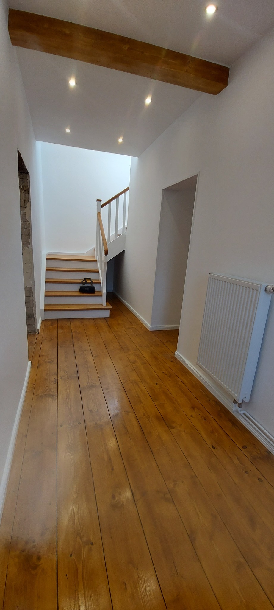 Jasne, odnowione drewniane deski podłogowe w korytarzu z białymi ścianami, widoczne schody z drewnianą balustradą i grzejnik.