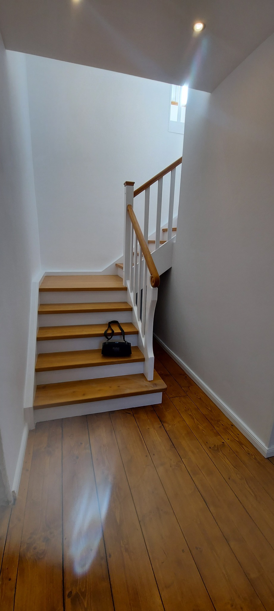 Jasne, drewniane schody z białymi poręczami i brązowym uchwytem, na trzecim stopniu leży czarna torba JBL, widoczne świeże białe ściany i drewniana podłoga.