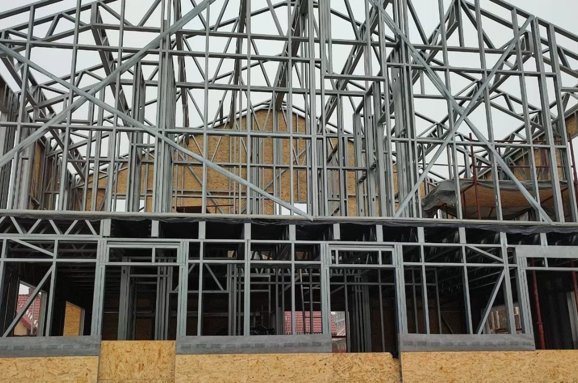 Konstrukcja szkieletowa domu w trakcie budowy, widoczne metalowe profile tworzące ściany i dach, płyty OSB wewnątrz konstrukcji, szara pogoda w tle.