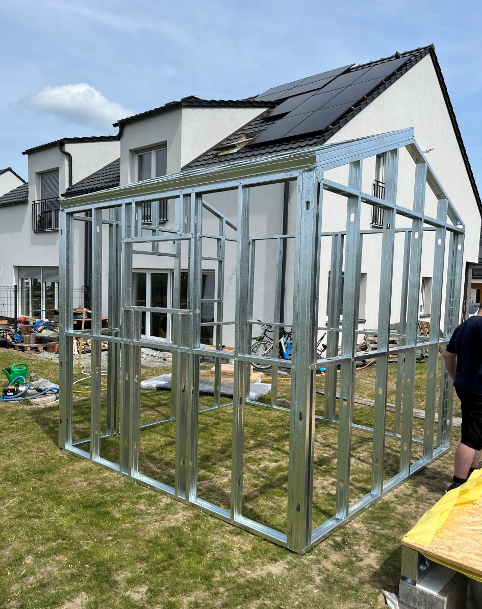 Metalowa konstrukcja szkieletowa domu jednorodzinnego na trawniku, widoczny fragment elewacji domu z panelami słonecznymi na dachu.