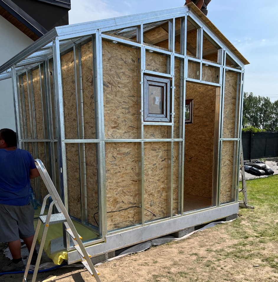 Konstrukcja szkieletowa budynku z płyt OSB i metalowych profili, widoczne okno z brązową ramą, drabina oparta o ścianę, fragment trawnika i ogrodzenia w tle.