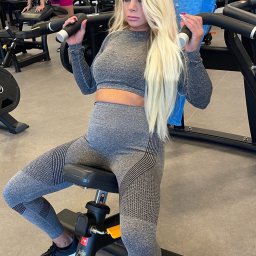 Wiktoria Wasik Trener Personalny - Kobieta w szarym sportowym stroju ćwiczy na maszynie w siłowni. Widoczne obciążenia, niebieski ręcznik i inne osoby w tle. Jasne włosy, profesjonalny makijaż.