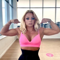 Wiktoria Wasik Trener Personalny - Kobieta w różowym topie prezentuje muskulaturę bicepsów w jasnym wnętrzu siłowni, z widocznym pasem wyszczuplającym talię i oknami w tle.