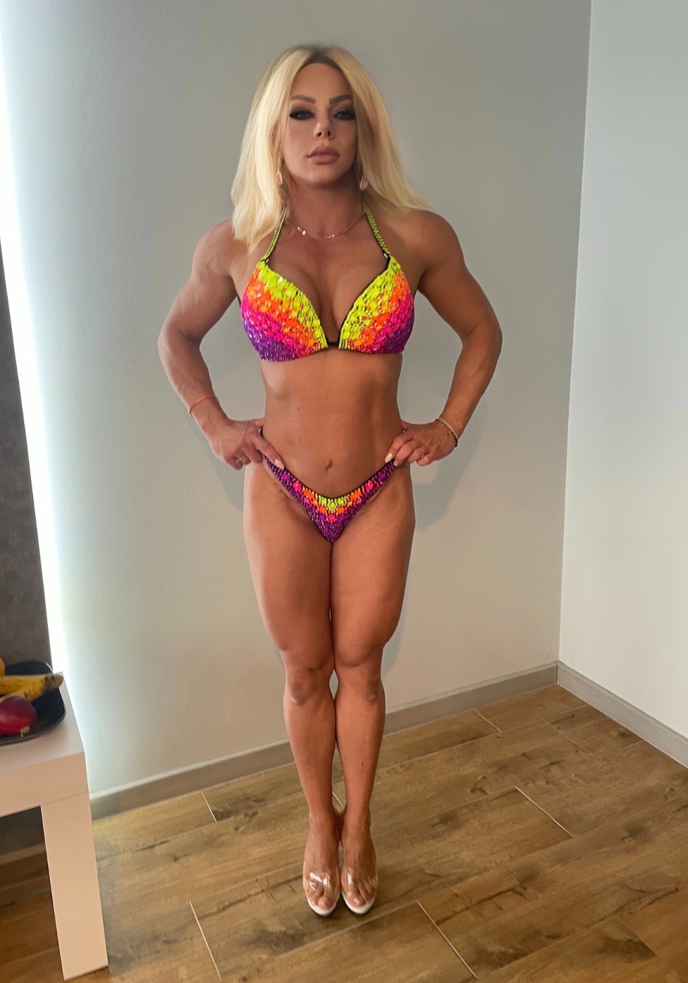 Kobieta o atletycznej budowie ciała prezentuje sylwetkę w bikini z cekinami w odcieniach neonowej żółci, pomarańczy i fioletu. Uwagę zwracają mocno zarysowane mięśnie.