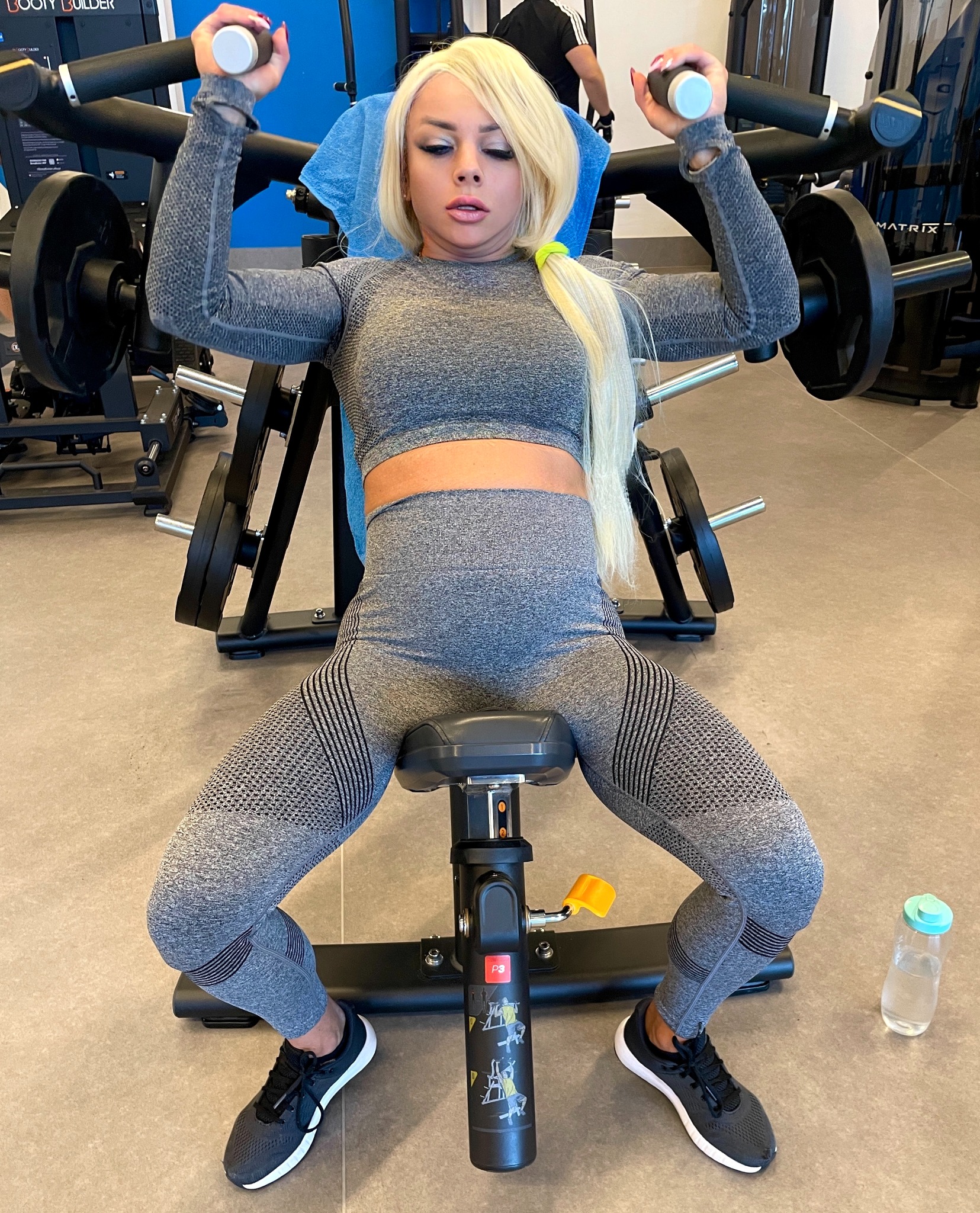 Blond kobieta w szarym stroju sportowym ćwiczy na maszynie do wyciskania w siłowni. Widoczna butelka wody i instrukcja obsługi na maszynie.