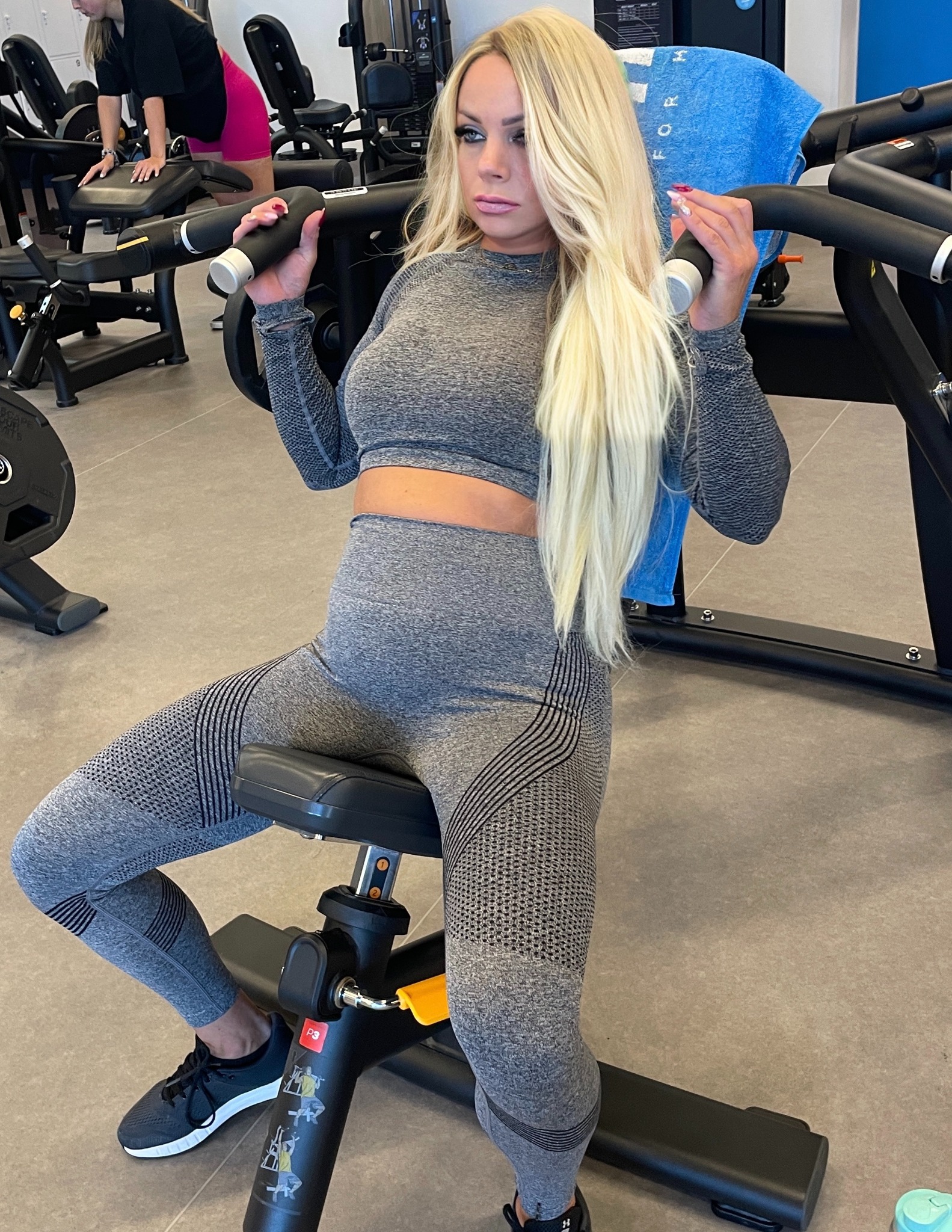 Kobieta w szarym sportowym stroju ćwiczy na maszynie w siłowni. Widoczne obciążenia, niebieski ręcznik i inne osoby w tle. Jasne włosy, profesjonalny makijaż.