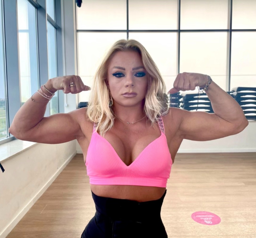 Kobieta w różowym topie prezentuje muskulaturę bicepsów w jasnym wnętrzu siłowni, z widocznym pasem wyszczuplającym talię i oknami w tle.