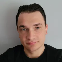 Portret młodego mężczyzny z ciemnymi włosami zaczesanymi do tyłu, ubrany w czarną koszulkę na tle szarej ściany, oświetlenie z góry.