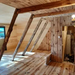 Poddasze z drewnianą konstrukcją więźby dachowej, drewnianą podłogą i boazerią ścienną w trakcie remontu, z widocznymi elementami oświetlenia i narzędziami w tle.