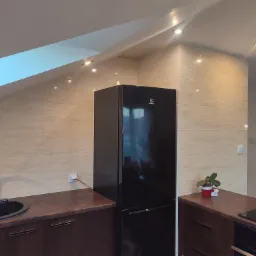 Wnętrze kuchni na poddaszu z beżowymi płytkami na ścianie, czarnym zlewem i czarną lodówką, oświetlone punktowymi lampami.