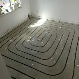 Szare rury ogrzewania podłogowego ułożone spiralnie w surowym, betonowym podłożu w pustym, jasnym pomieszczeniu, z widocznym fragmentem okna i roślinami doniczkowymi w tle.