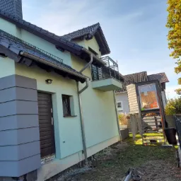 Jasnozielony dom jednorodzinny z ciemnoszarą elewacją, balkonem z metalową balustradą i ciemnym dachem z dachówki, widoczny z niskiej perspektywy.