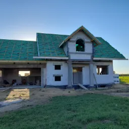 Surowy stan domu jednorodzinnego z białych bloczków, z zieloną membraną dachową i drewnianą konstrukcją dachu, w słoneczny dzień.