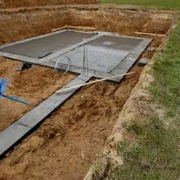 Wykop pod fundamenty z wylanym betonem, widoczne zbrojenie, szalunki i narzędzia w wykopie z ziemi.