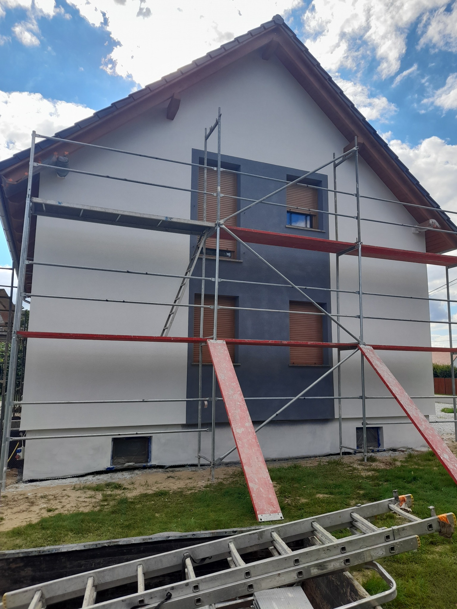 Dom jednorodzinny w trakcie prac elewacyjnych, widoczne rusztowanie, fragment pomalowanej ściany na szaro i biało, zabezpieczone okna.