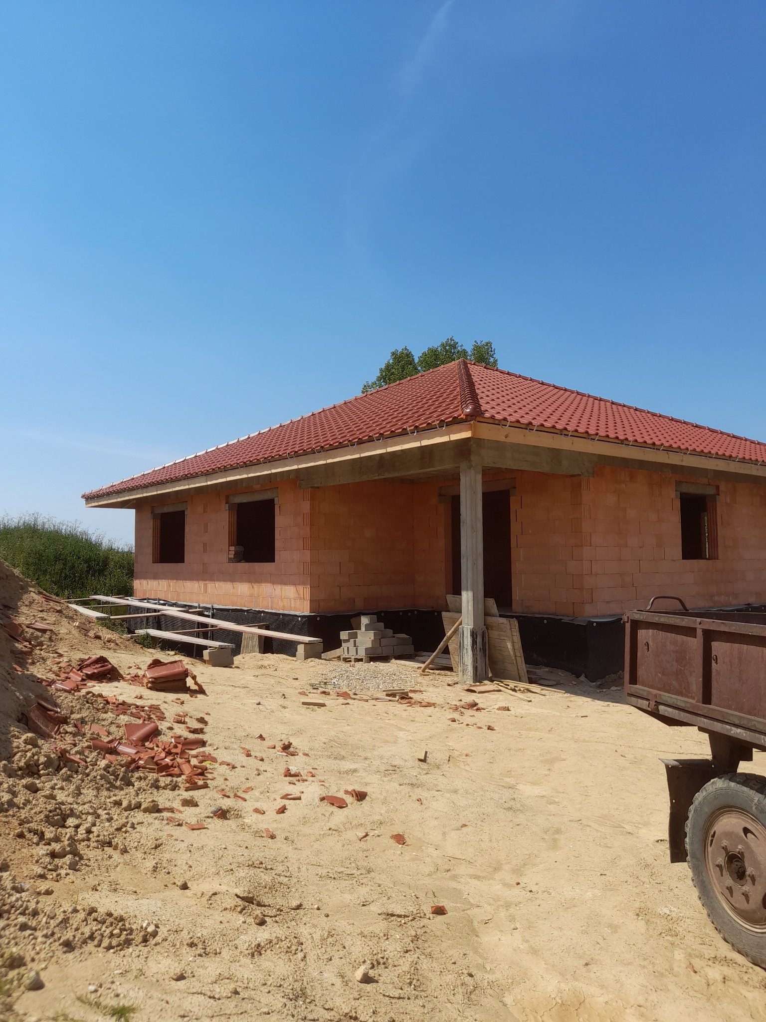 Niedokończony dom jednorodzinny z czerwonej cegły ceramicznej i czerwoną dachówką, widoczne fundamenty i fragment przyczepy na piaszczystym terenie budowy.