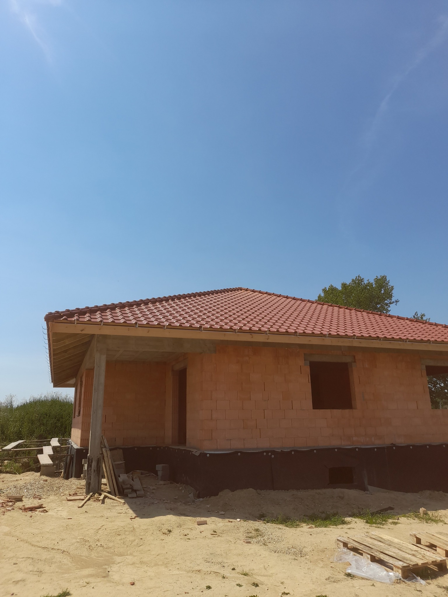 Budowa domu jednorodzinnego w stanie surowym otwartym: murowane ściany z czerwonej cegły, czerwona dachówka, widoczne fundamenty z izolacją, niezagospodarowany teren wokół.