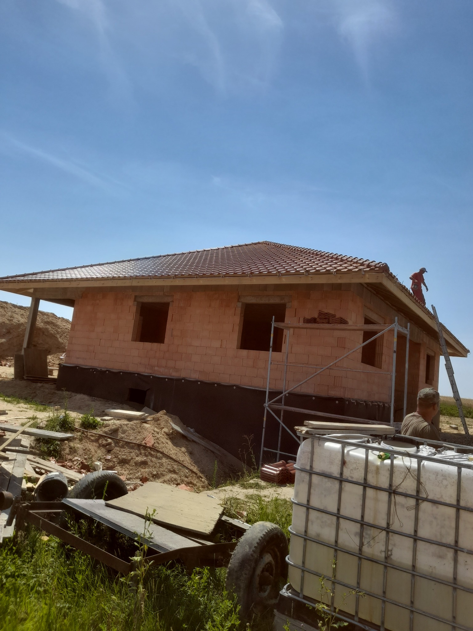 Surowy stan budynku jednorodzinnego z czerwonej cegły z zamontowaną częścią dachu z czerwonej dachówki, widoczne rusztowania, mężczyzna na dachu w czerwonym kombinezonie, zbiornik na wodę...