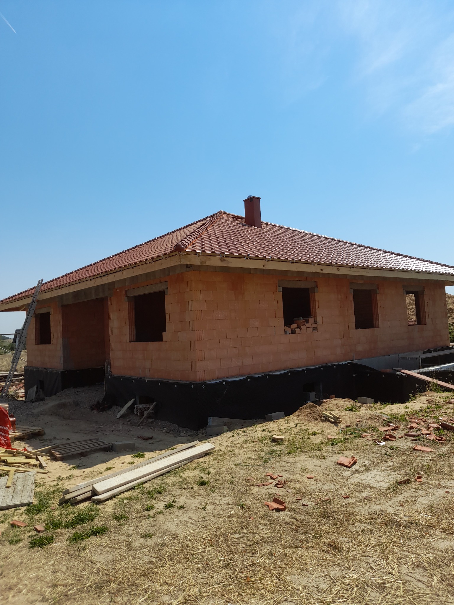 Surowy stan domu jednorodzinnego z czerwonej cegły z dachem z dachówki ceramicznej w trakcie budowy, widoczne fragmenty fundamentów i rusztowania.