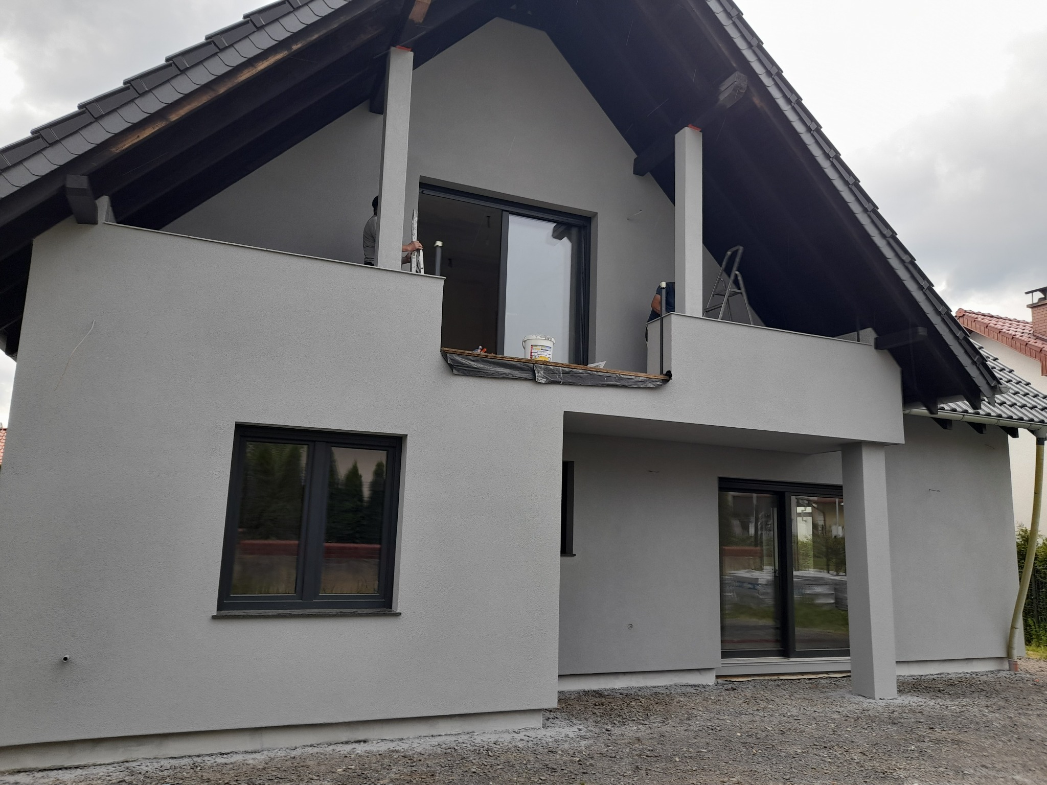 Dom jednorodzinny z szarą elewacją w trakcie prac wykończeniowych, widoczni pracownicy na balkonie, drabina i wiadro z farbą.