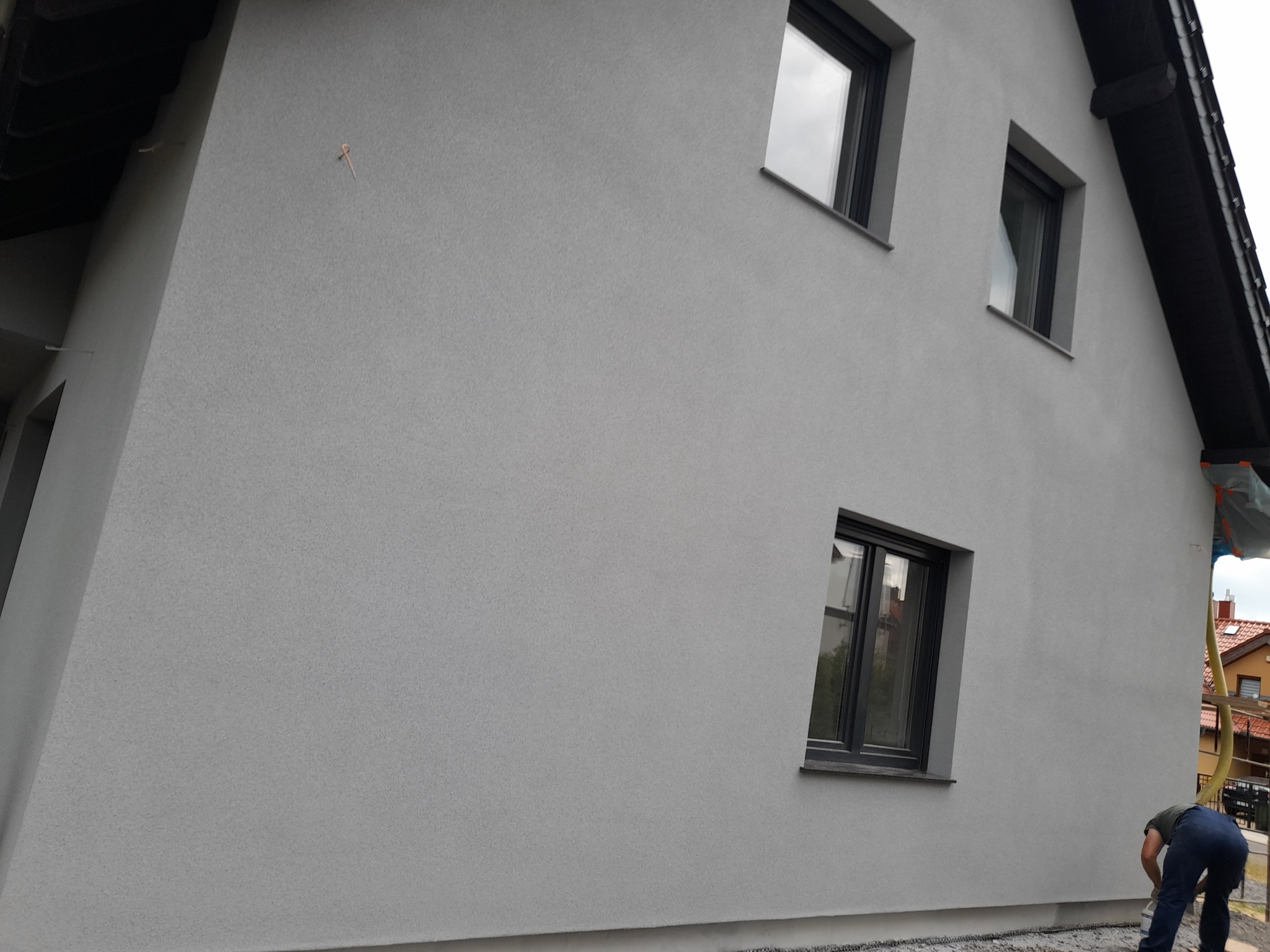 Świeżo otynkowana elewacja budynku jednorodzinnego w odcieniach szarości, widoczne trzy okna z ciemnymi ramami, osoba w dolnym rogu przy podstawie ściany.