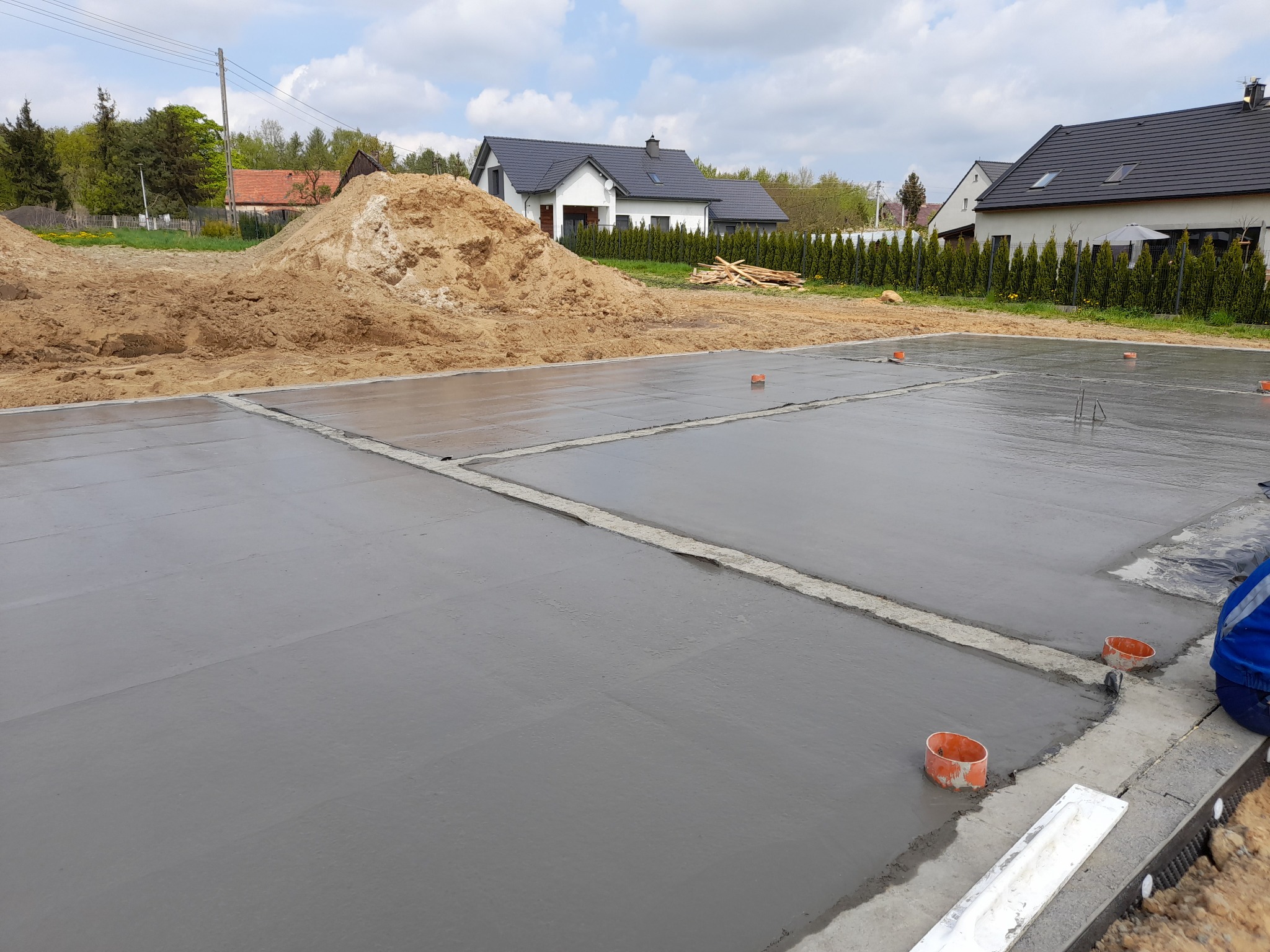 Wylana, świeża płyta fundamentowa z widocznymi dylatacjami i pomarańczowymi rurkami, w tle hałda piachu i domy jednorodzinne pod błękitnym niebem.
