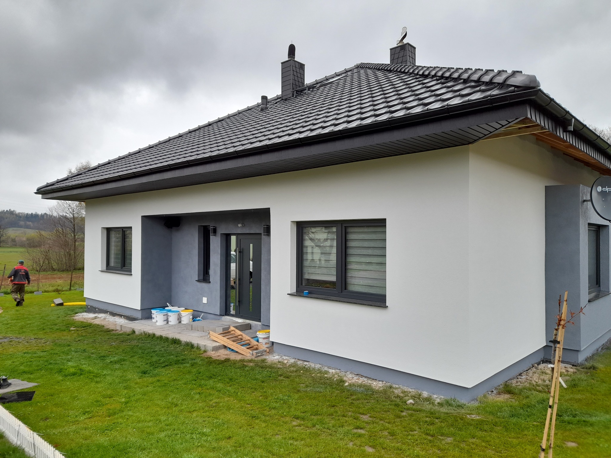 Świeżo otynkowana elewacja domu jednorodzinnego z ciemną stolarką okienną i drzwiami, widoczne wiaderka z farbą i drabinka, pracownik w oddali.