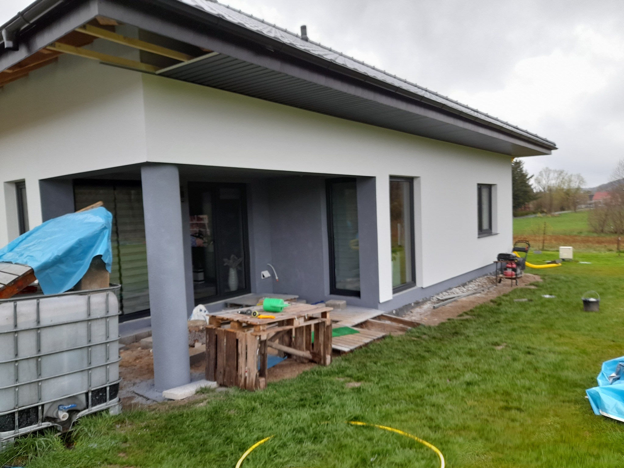 Nowoczesny dom jednorodzinny z szarą stolarką okienną po świeżym pomalowaniu elewacji na biało, widoczne tymczasowe podesty i narzędzia przygotowane do dalszych prac wykończeniowych.