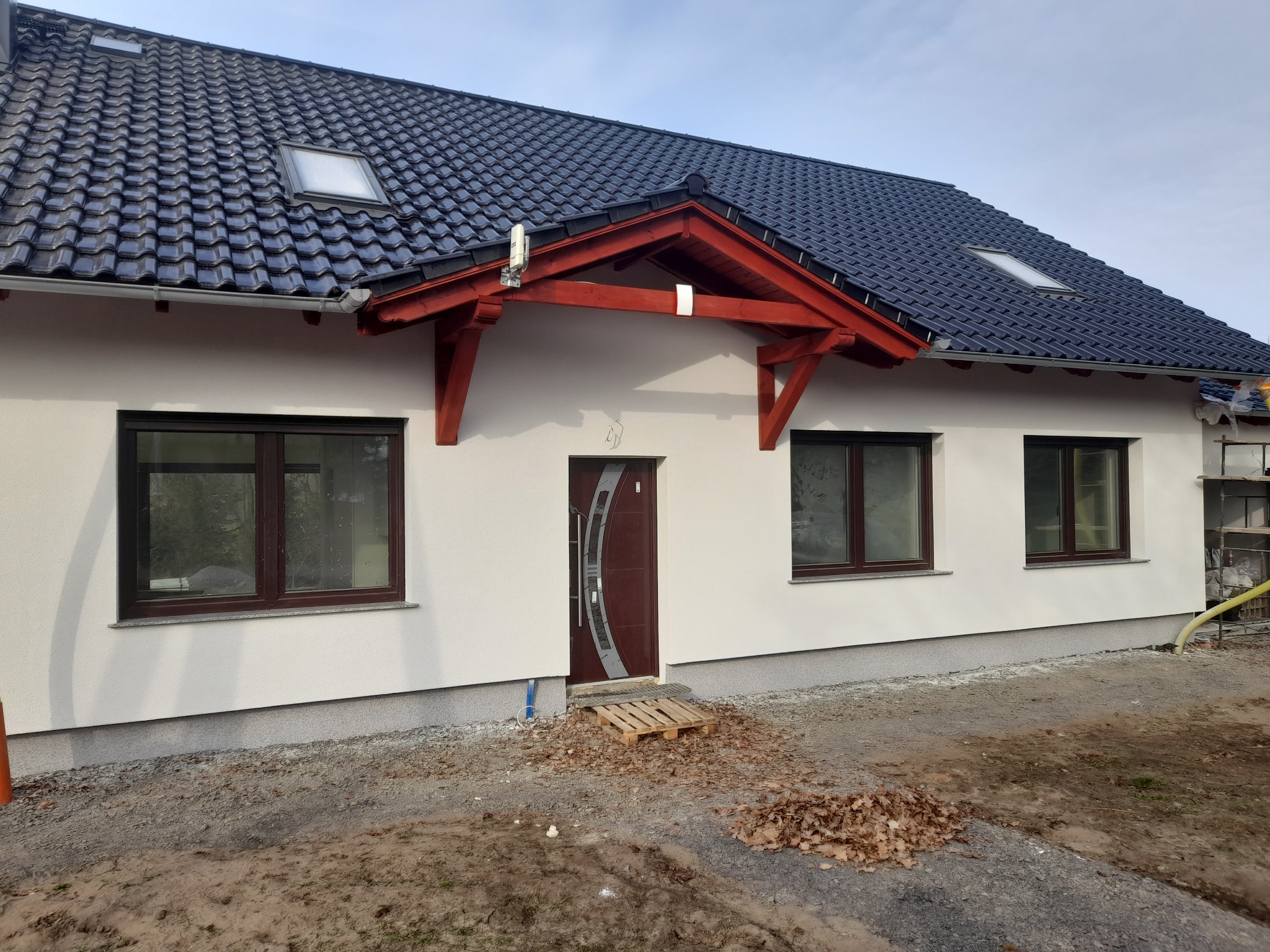 Nowy dom jednorodzinny z białą elewacją, ciemnobrązowymi oknami i drzwiami, oraz dachem z ciemnej dachówki, widoczne elementy wykończeniowe fundamentów, podbitka dachowa w kolorze czerwonym, teren...