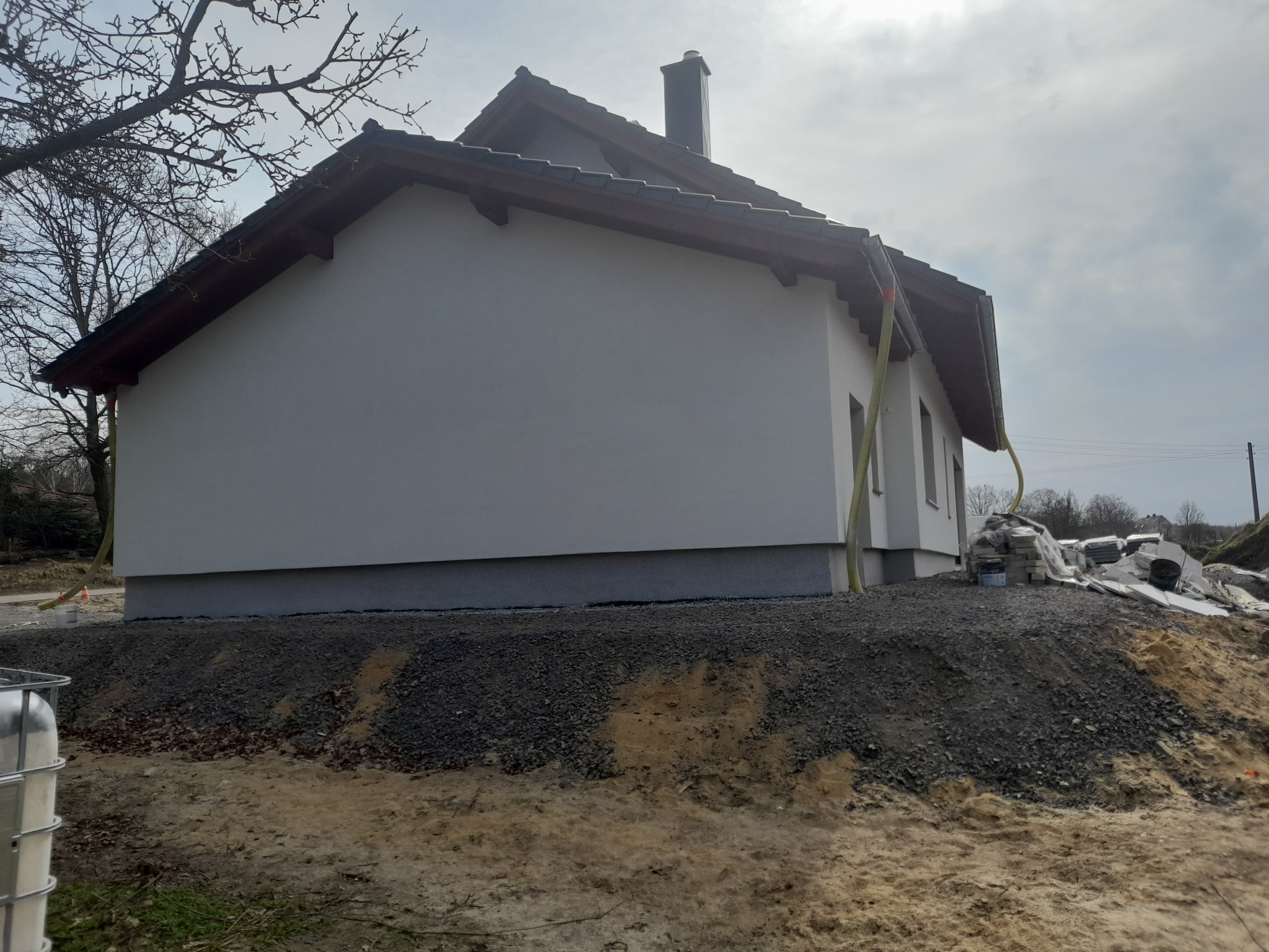 Świeżo otynkowana ściana domu jednorodzinnego z widocznym fragmentem dachu i komina, teren wokół domu w trakcie wyrównywania, widoczne materiały budowlane i wąż do tynkowania.