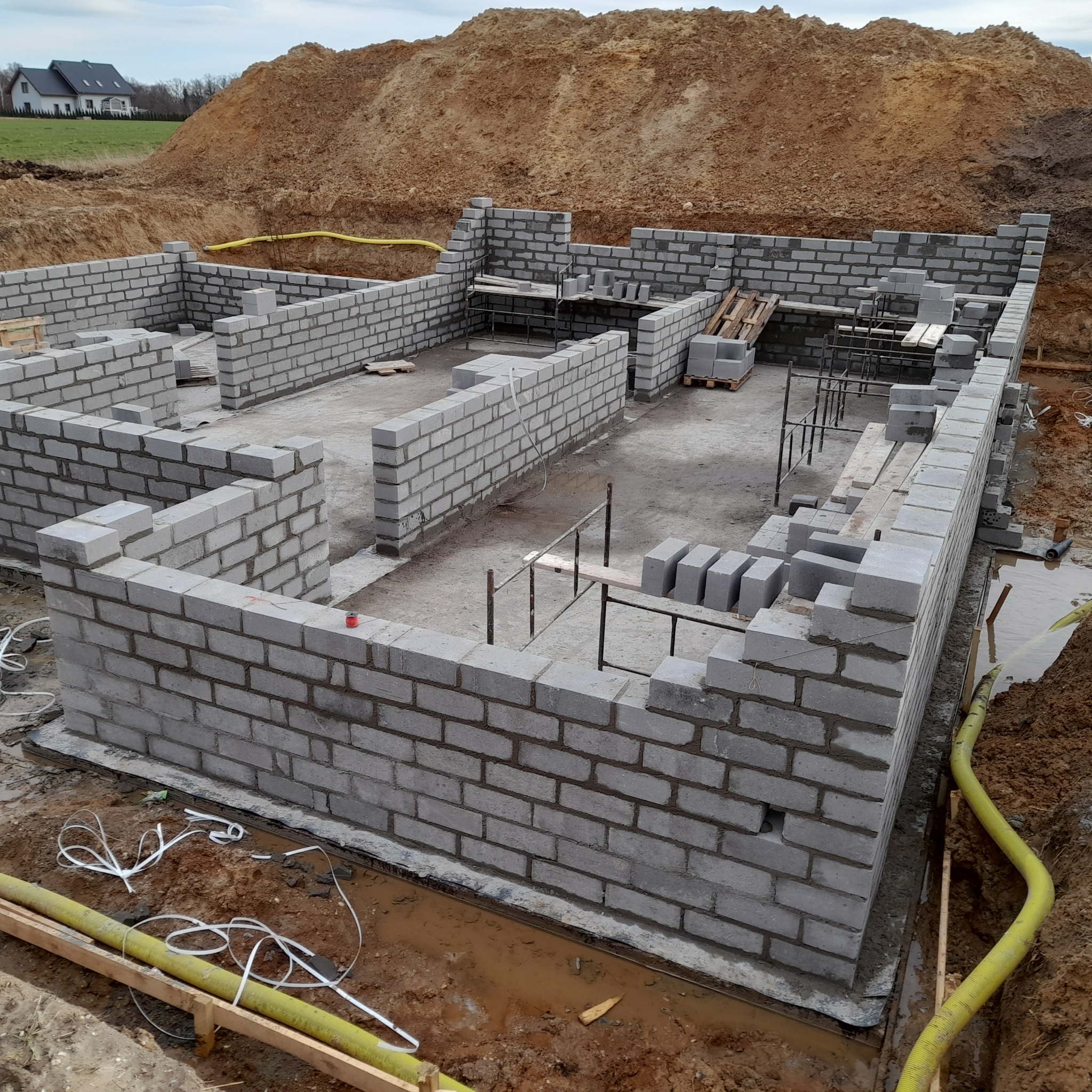 Wznoszenie ścian fundamentowych z bloczków betonowych, widoczne zbrojenie i żółty wąż odprowadzający wodę, w tle hałda ziemi i dom.