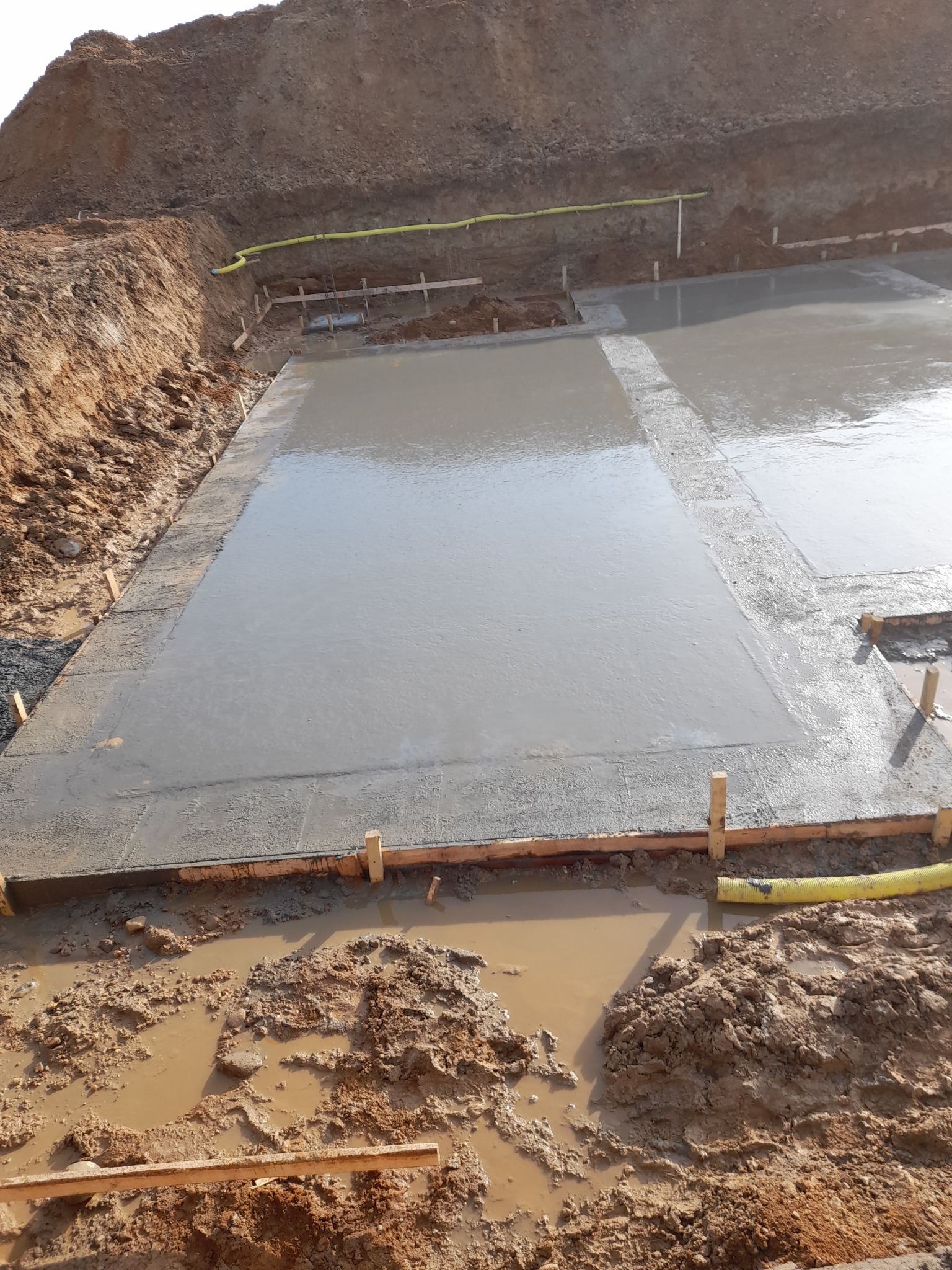 Świeżo wylana, mokra płyta fundamentowa w drewnianym szalunku, z widocznym żółtym wężem doprowadzającym wodę i błotnistym terenem dookoła.