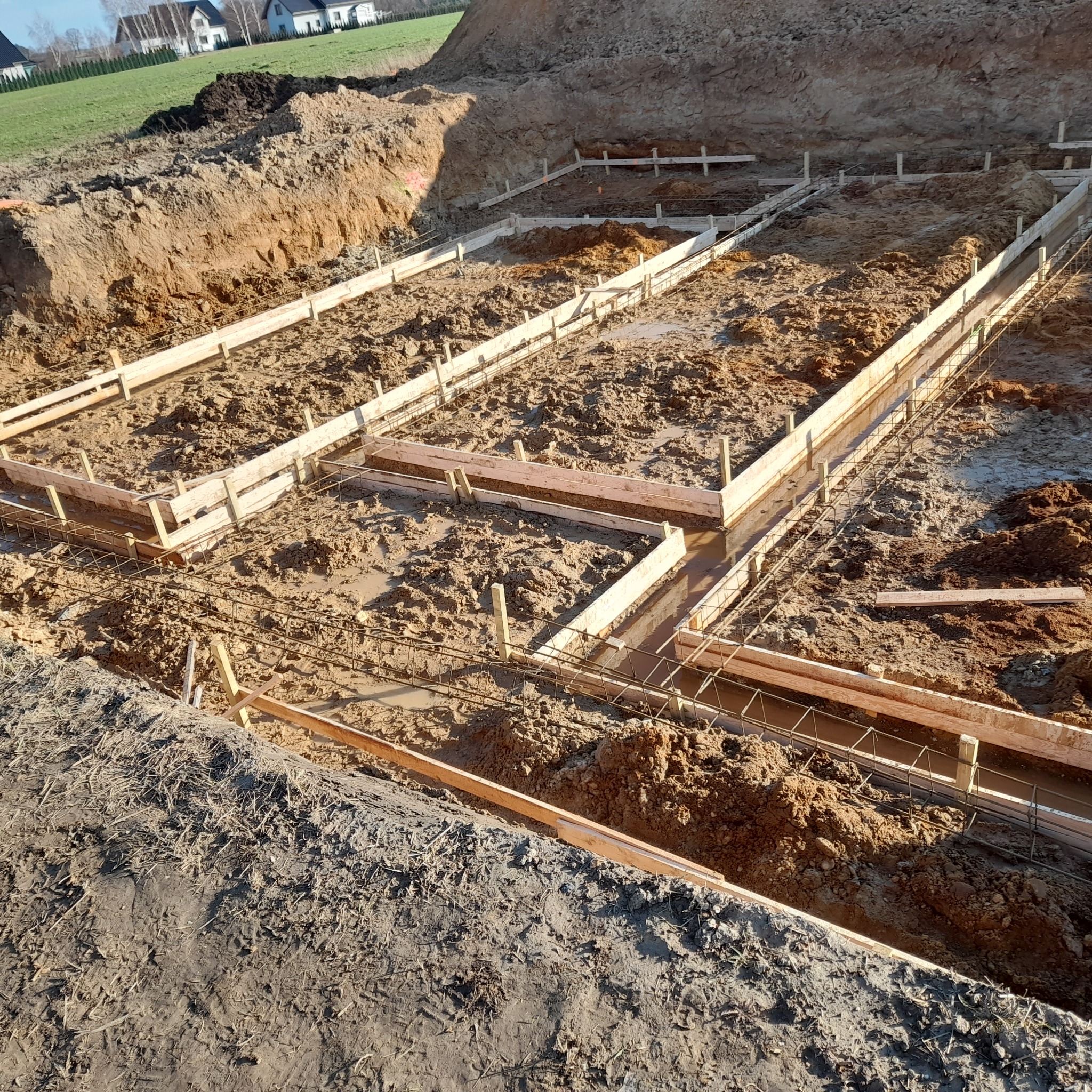 Przygotowanie wykopu pod fundamenty z drewnianym szalunkiem i zbrojeniem stalowym, widoczne ślady wody i błota w wykopie, w tle zabudowa jednorodzinna.