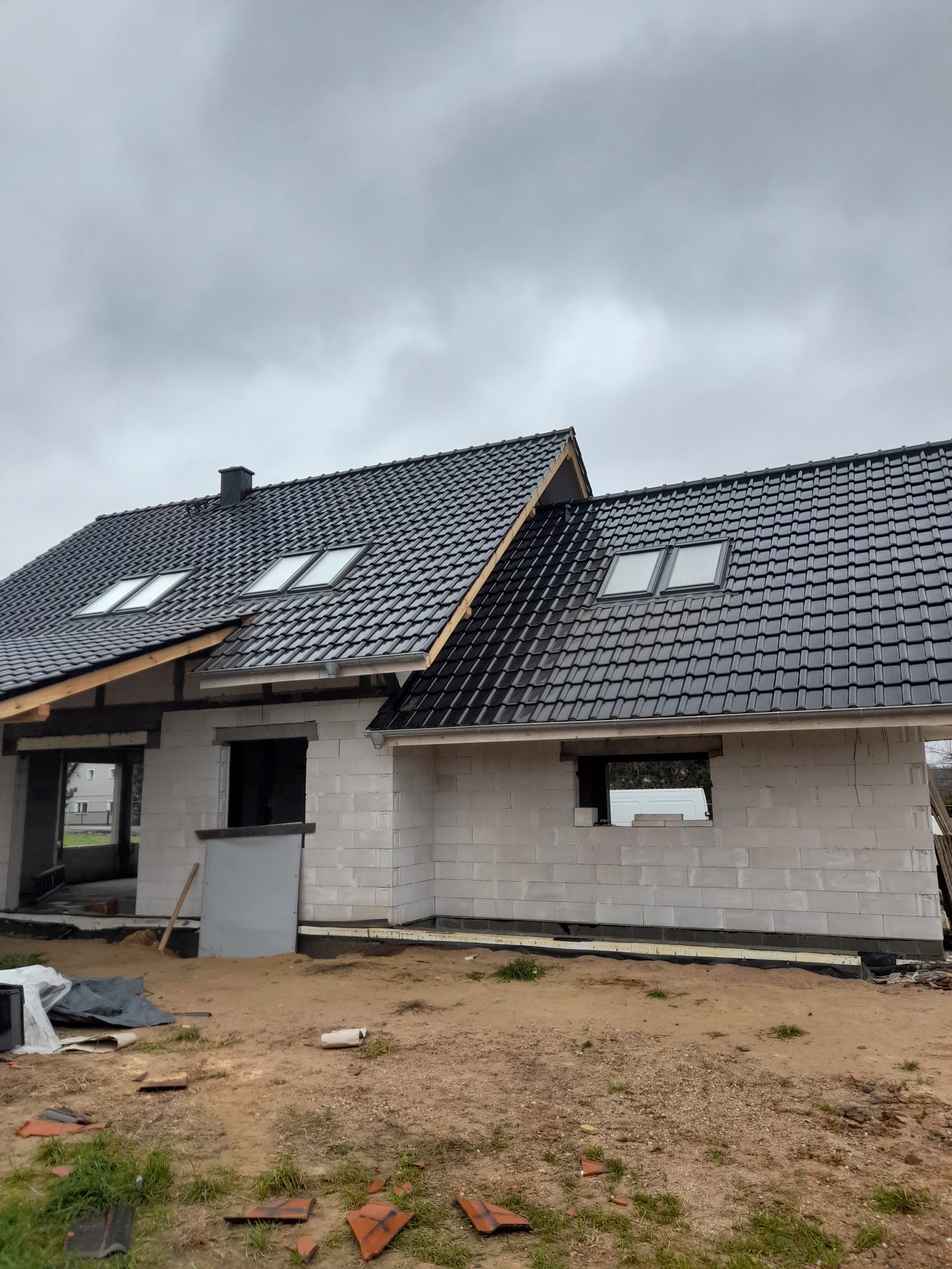 Budowa domu jednorodzinnego w stanie surowym otwartym: ściany z bloczków, czarny dach z oknami dachowymi, widoczne fragmenty fundamentów i izolacji przeciwwilgociowej.