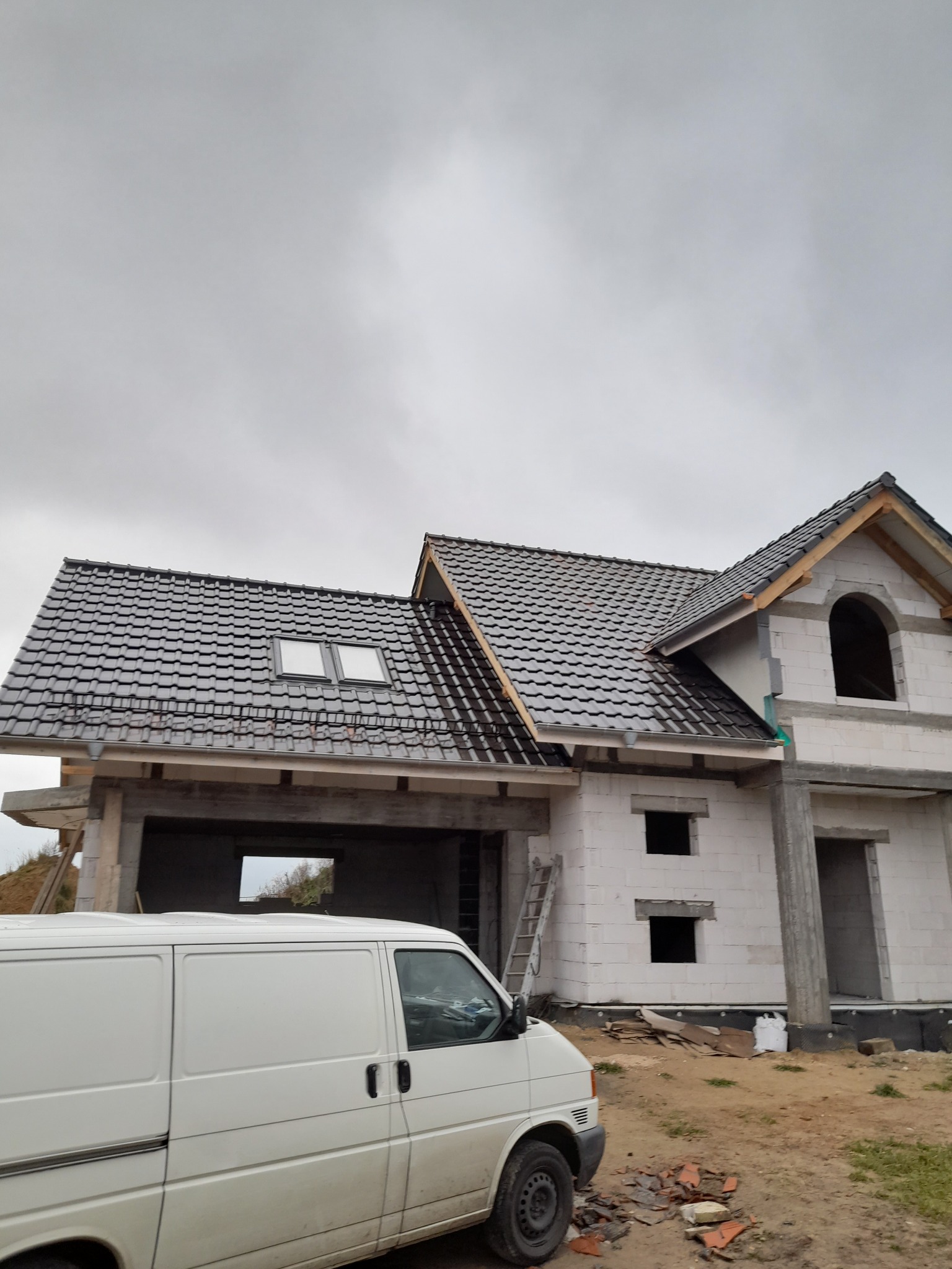 Niedokończony dom z białych bloczków, widoczny fragment dachu z dachówką, biały samochód dostawczy na pierwszym planie, ziemiste podłoże i pochmurne niebo.