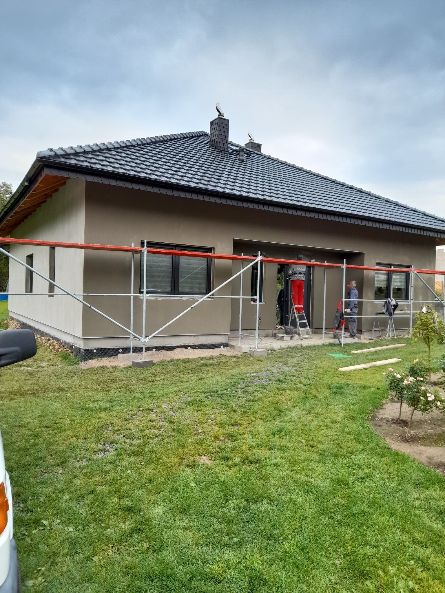 Dom jednorodzinny w trakcie prac wykończeniowych elewacji w Bolesławcu, widoczne rusztowania, pracownicy przy wejściu, szara, świeżo nałożona warstwa tynku.