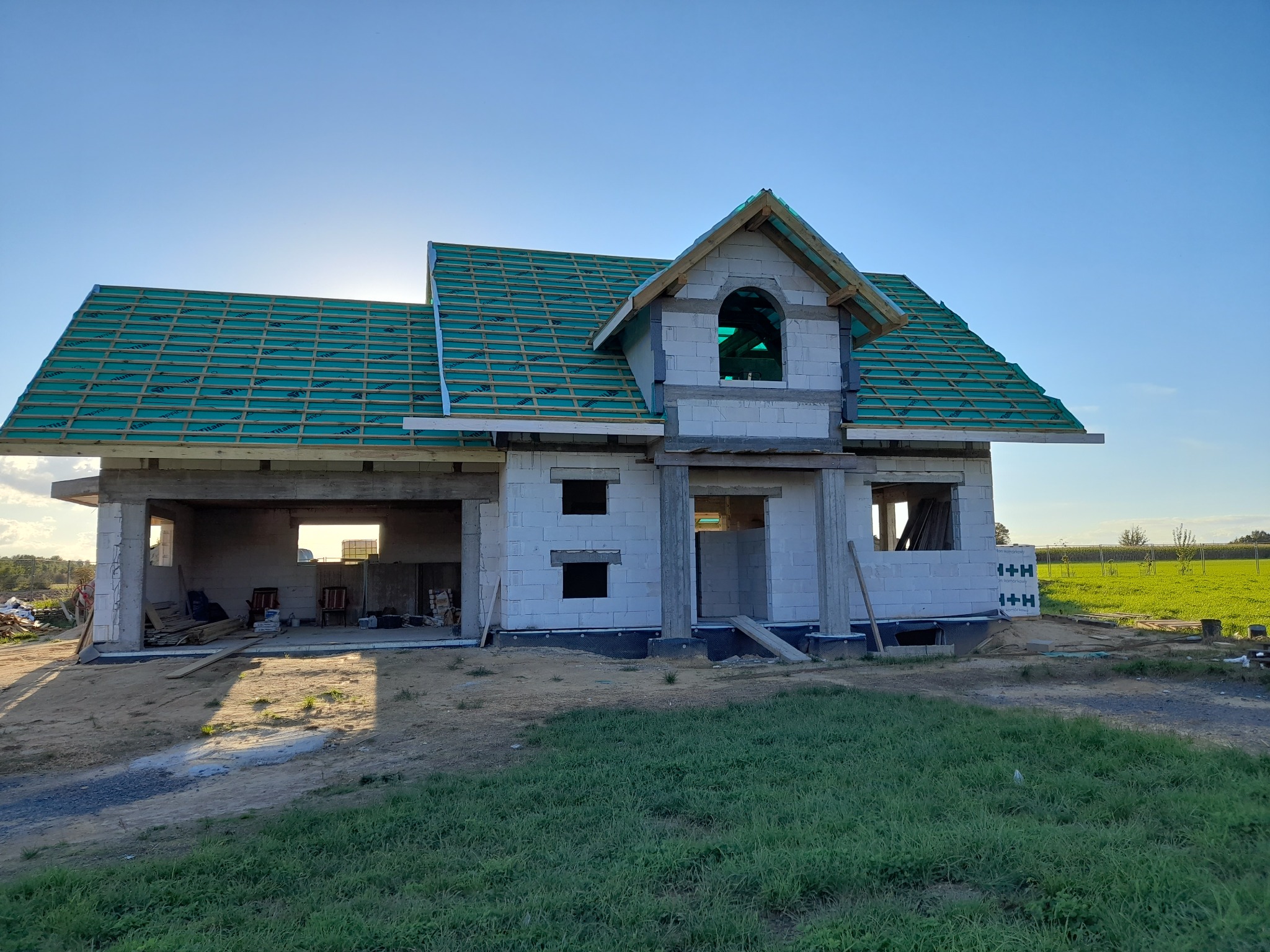Surowy stan domu jednorodzinnego z białych bloczków, z zieloną membraną dachową i drewnianą konstrukcją dachu, w słoneczny dzień.