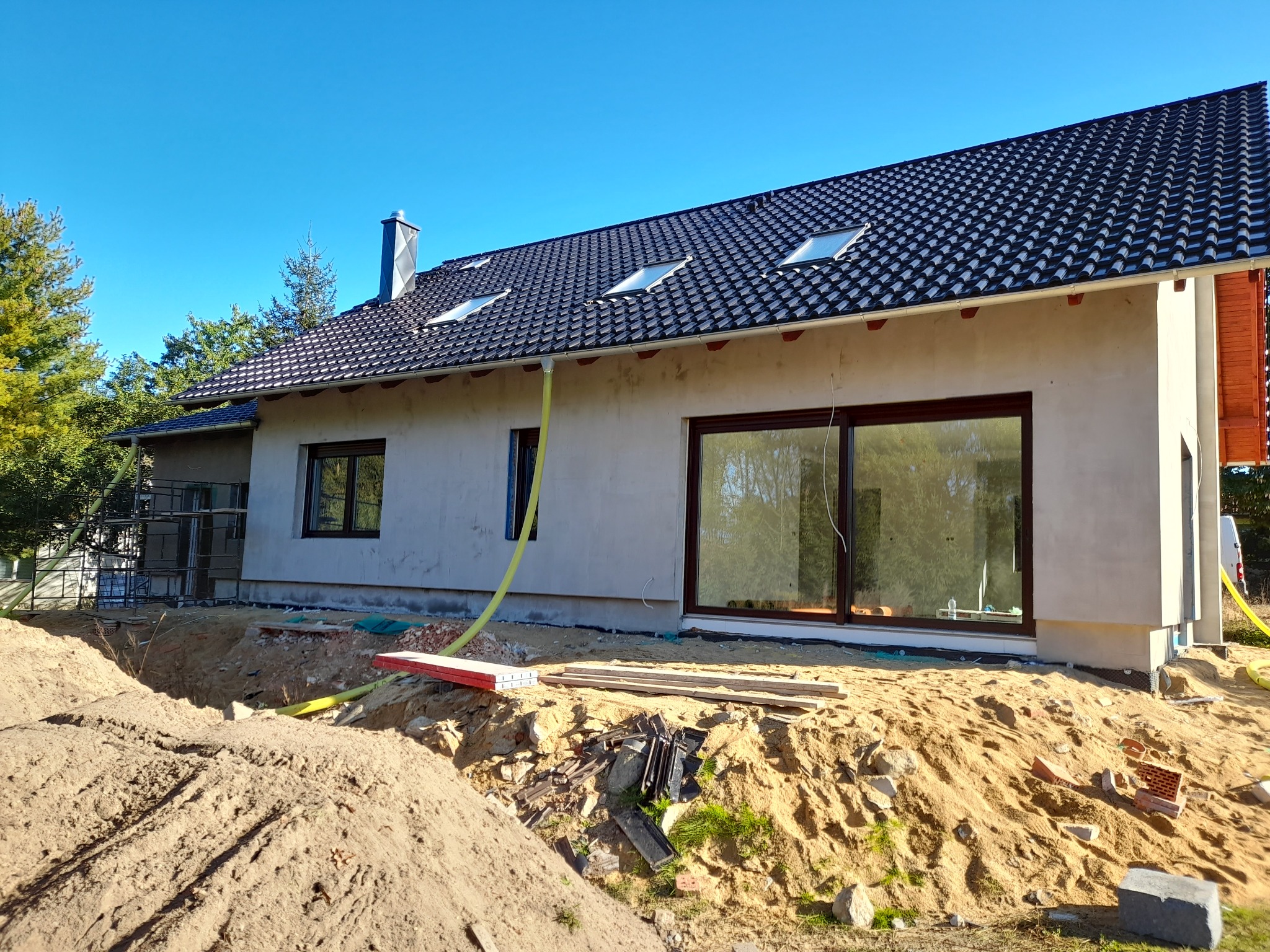 Dom jednorodzinny w trakcie ocieplania elewacji, widoczne rusztowania, stos piasku i przygotowane materiały budowlane, dach pokryty czarną dachówką z oknami dachowymi, widoczny fragment elewacji...