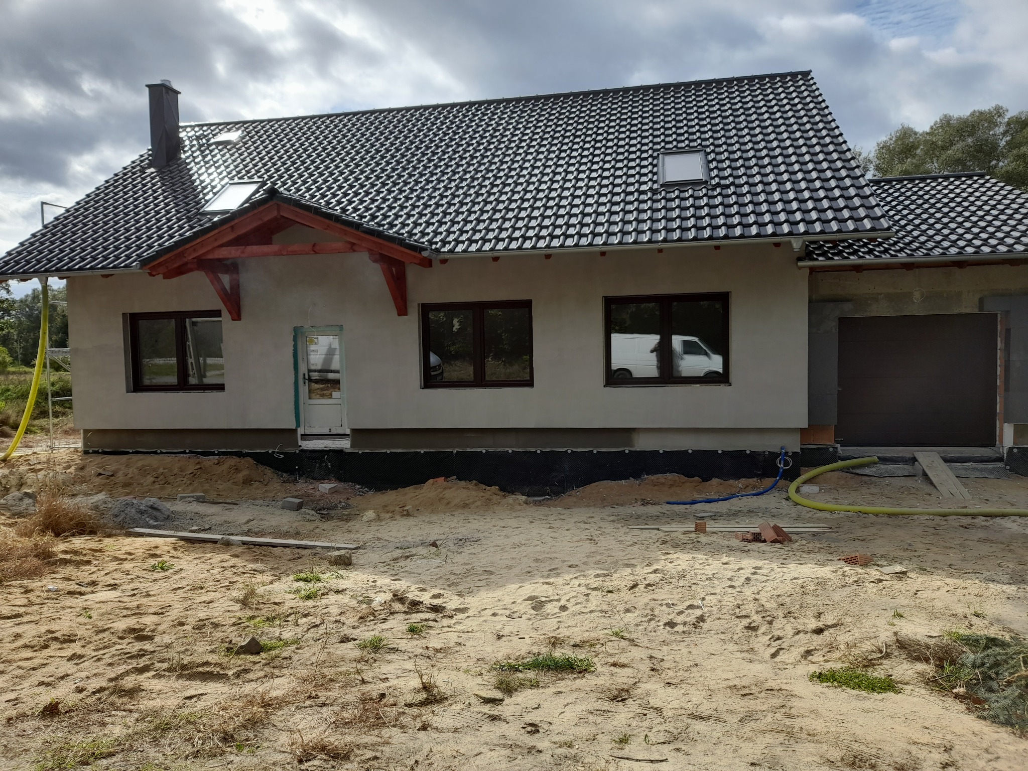Surowy stan elewacji domu jednorodzinnego z czarną dachówką, widoczne okna, drzwi wejściowe i brama garażowa, teren wokół w trakcie przygotowania.