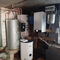 N-Energia - Instalacja grzewcza w piwnicy z kilkoma zbiornikami, kotłem i miedzianymi rurami, widok na okno i surowy betonowy strop.