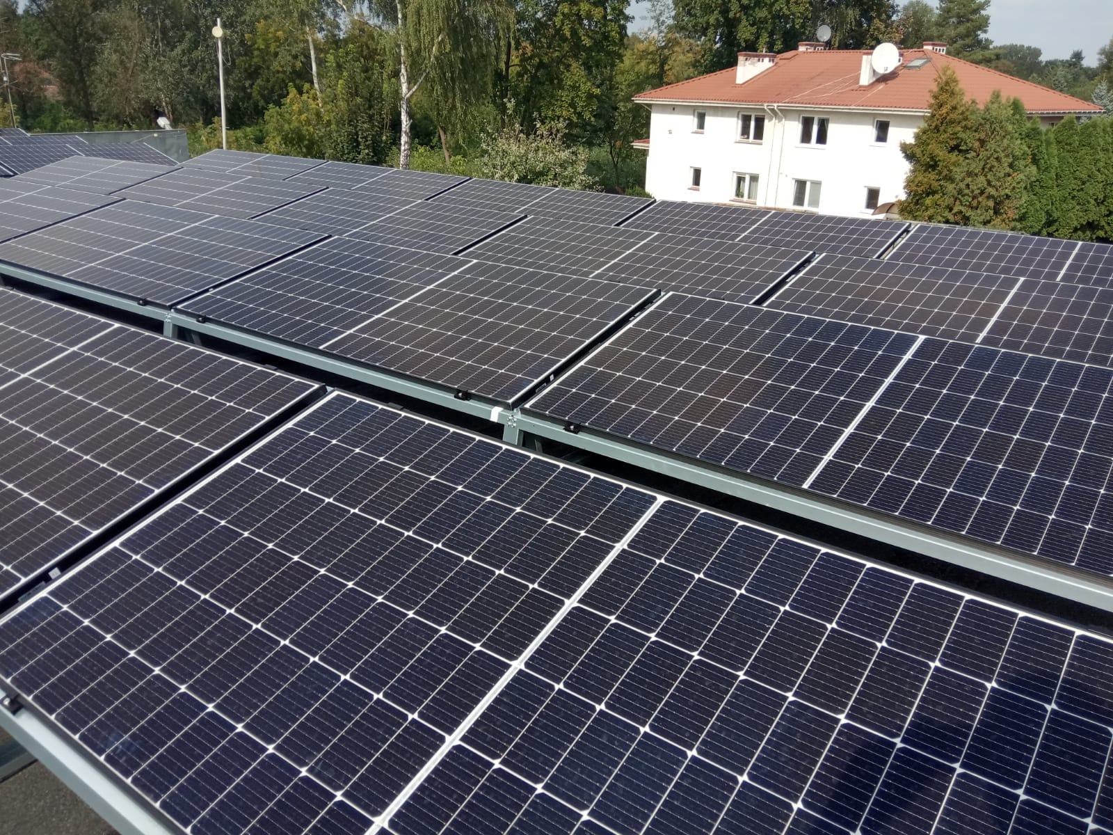 Rząd paneli słonecznych na dachu z widokiem na biały, dwupiętrowy dom z czerwoną dachówką i zielonymi drzewami w tle.