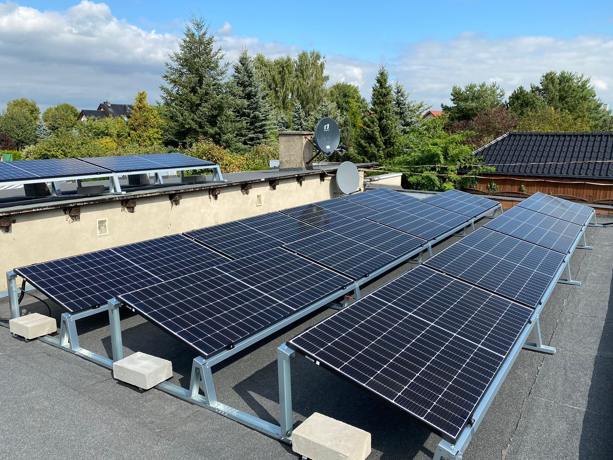Rząd paneli słonecznych na płaskim dachu, wspartych na metalowej konstrukcji z betonowymi obciążnikami, w tle drzewa i antena satelitarna.