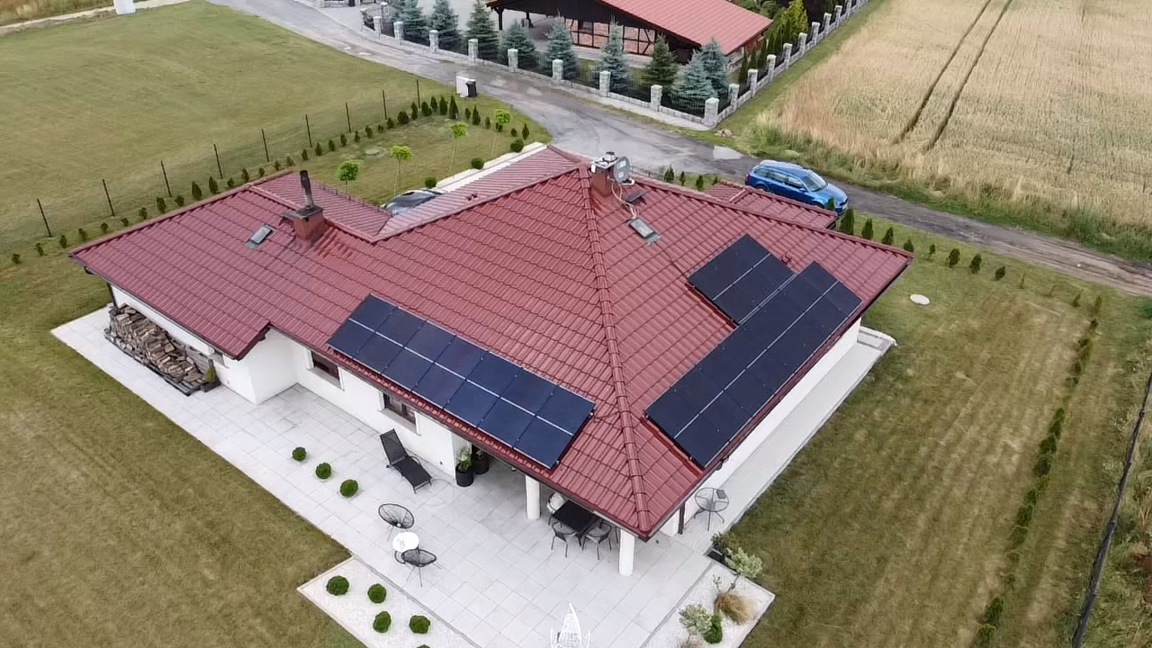 Widok z lotu ptaka na dom z czerwoną dachówką i panelami słonecznymi na dachu, otoczony zielonym trawnikiem i drzewami.