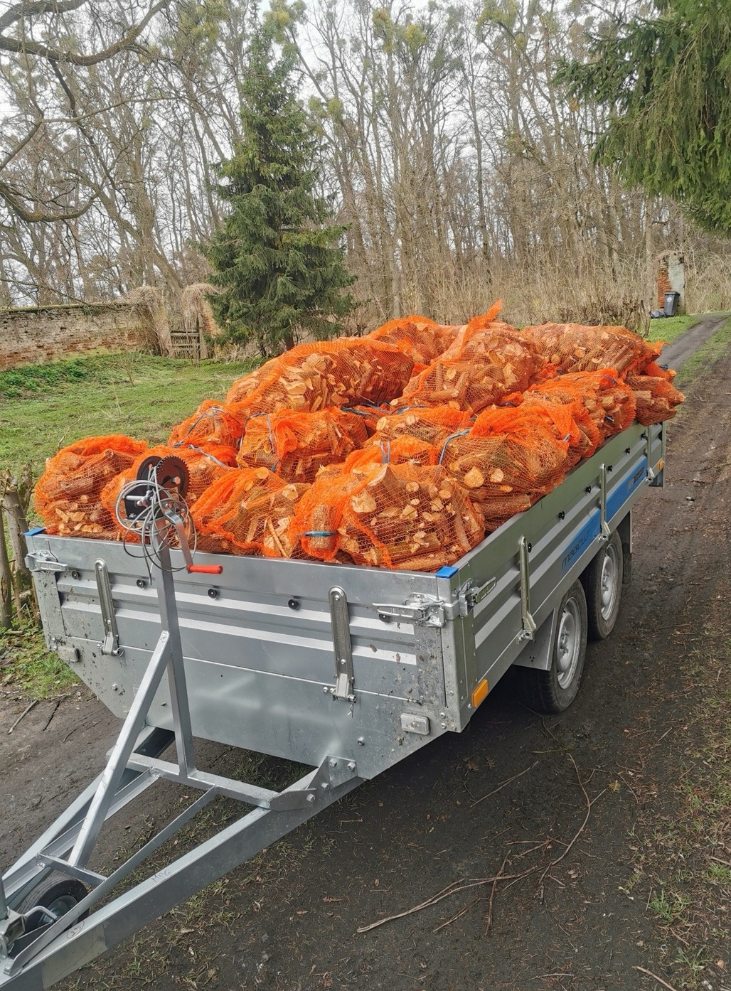 Przyczepa załadowana workami pomarańczowej siatki wypełnionymi pociętym drewnem kominkowym, na tle drzew i zarośli.