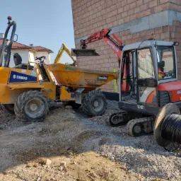 Żółta wywrotka Thwaites i czerwona minikoparka Kubota obok fundamentów budynku z cegły, na pierwszym planie bęben kablowy.