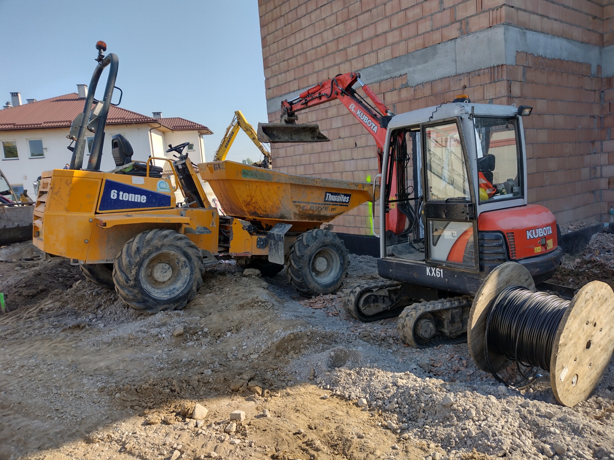 Żółta wywrotka Thwaites i czerwona minikoparka Kubota obok fundamentów budynku z cegły, na pierwszym planie bęben kablowy.